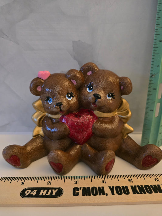 Two small brown teddy bears holding a red heart with a ruler for scale on a white background